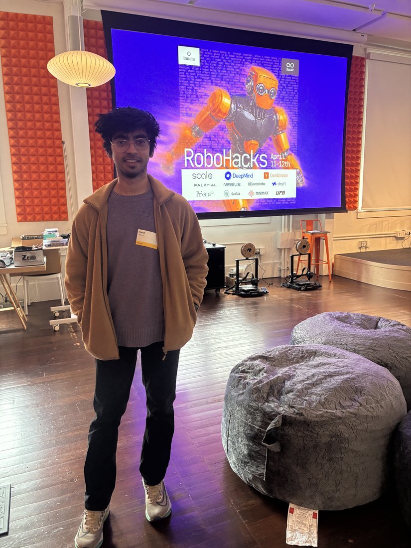 Harsh Sharma at RoboHacks, standing in front of the event screen showing the RoboHacks robot artwork and sponsor logos including Scale, Google DeepMind, Y Combinator, Nebius and ElevenLabs.